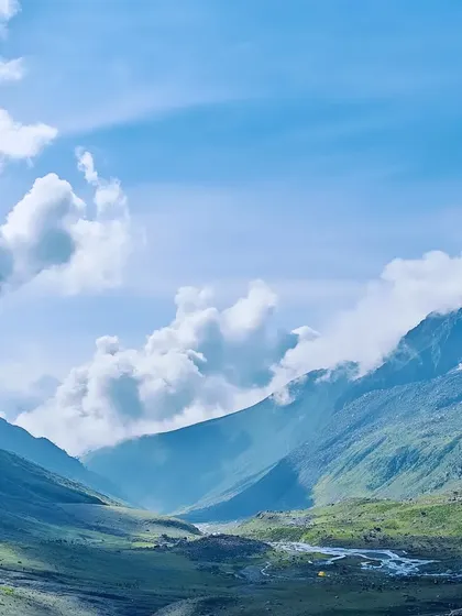 A panoramic view of a valley in Kashmir, where a river winds through lush green fields under a sky filled with soft, white clouds.