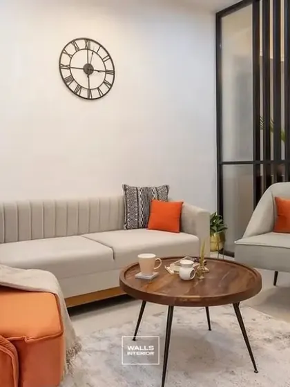 A comfortable living room setting with a mix of neutral and bold orange seating. The minimalist wall and large clock create a clean backdrop, while the wooden partition adds a touch of warmth and texture.