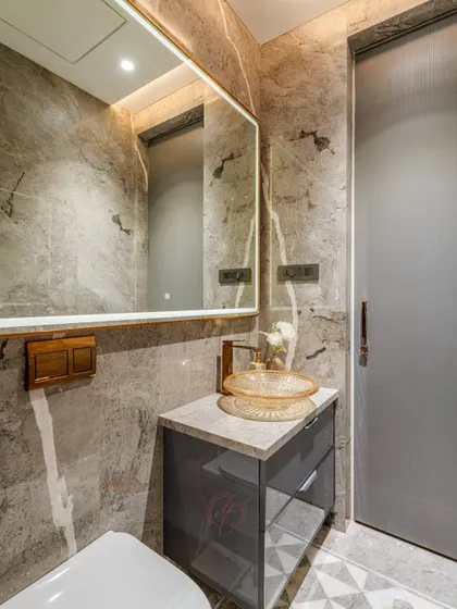 This bathroom uses a sophisticated palette of grey marble with brushed gold fixtures. The textured glass basin and large, back-lit mirror enhance both the aesthetics and the utility of the space.