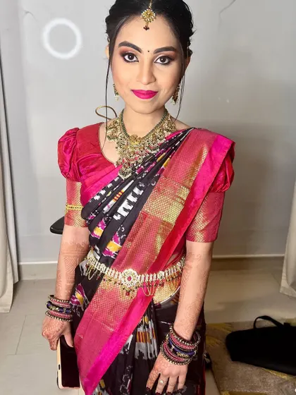 A full portrait of Bhanu, showcasing her complete bridal look. She wears a unique black and pink silk saree, complemented by antique-style gold jewellery and a pop of bright pink on her lips.