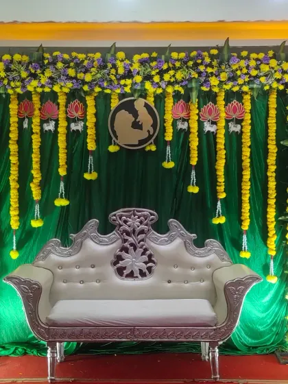 A traditional baby shower stage featuring a vibrant green backdrop with hanging yellow marigold garlands and lotus flower cutouts.