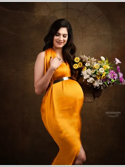 A soft and gentle portrait against a classic brown backdrop. The client, in a mustard gown, holds a basket of flowers, creating a warm and timeless maternity photo.