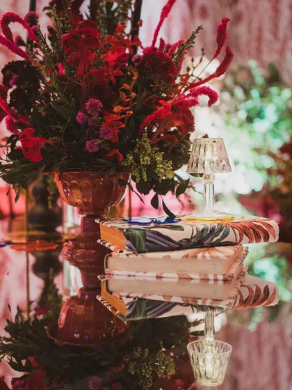 A close-up of a tabletop detail, featuring a vibrant red floral arrangement, a stack of custom-covered books, and a tiny, elegant table lamp.