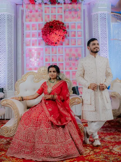 A regal portrait of the couple at their reception. The bride is seated on a throne-like chair while the groom stands beside her, creating a classic and elegant wedding portrait.