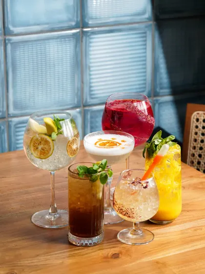 A vibrant collection of five different cocktails and coolers, expertly styled on a wooden table. This image is perfect for a bar or restaurant menu, showcasing the colorful and diverse drink offerings.