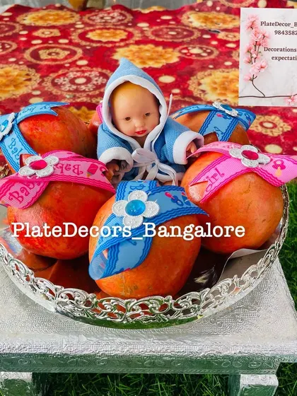 A creative baby shower idea where pomegranates are decorated with 'Baby Boy' and 'Baby Girl' ribbons. A doll sits in the middle of this unique fruit arrangement.