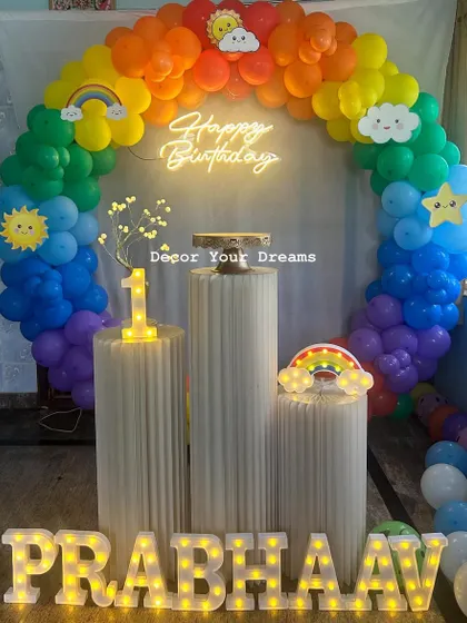 A cheerful rainbow-themed first birthday party. The decoration includes a full rainbow balloon arch, sun and cloud cutouts, and a light-up number '1'.
