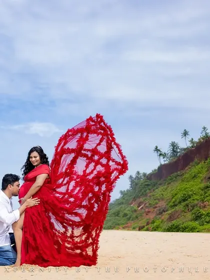 A tender moment as the father-to-be kisses the baby bump, with the dramatic red gown flowing around them.