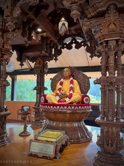 A life-like statue of a spiritual guru seated in a meditative pose within a hall supported by intricately carved wooden pillars.
