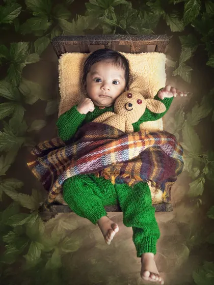 A creative composite shot of a two-month-old baby tucked into a tiny bed, surrounded by a frame of green leaves, creating a cozy, nest-like feel.