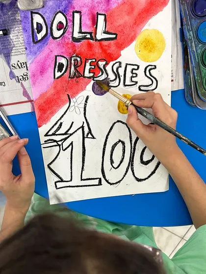 "Doll Dresses 100." A young artist creates a vibrant poster to advertise her handmade products for the school flea market.