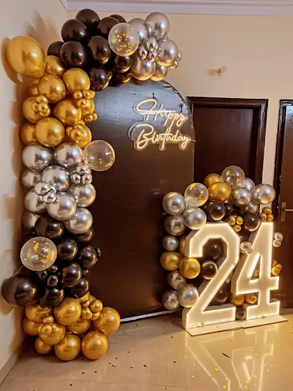 A bold and stylish 24th birthday decoration. The setup includes a black arched backdrop, a gold, black, and silver balloon garland, a neon sign, and marquee numbers.