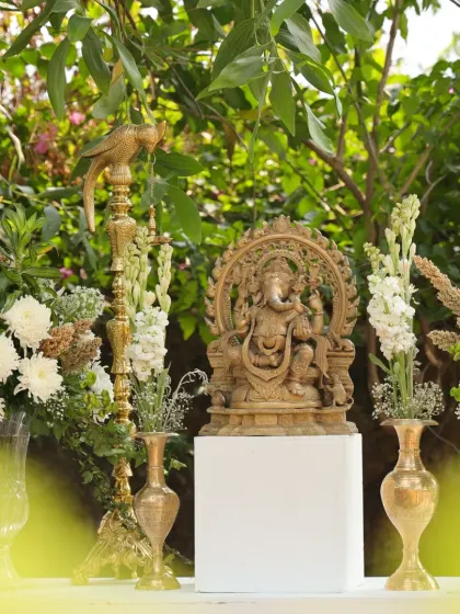 A beautifully carved wooden Ganesha statue, placed on a pedestal and surrounded by arrangements of white tuberoses and other flowers.