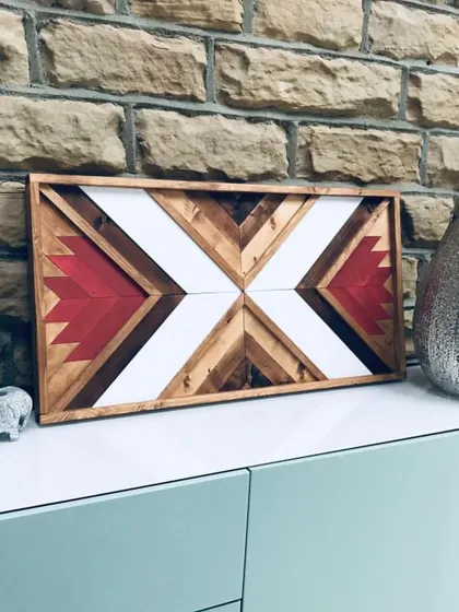This large Aztec wood wall art piece features a striking pattern in red, white, and various wood tones. It's displayed here on a console against a stone wall, showcasing its rustic yet modern appeal.