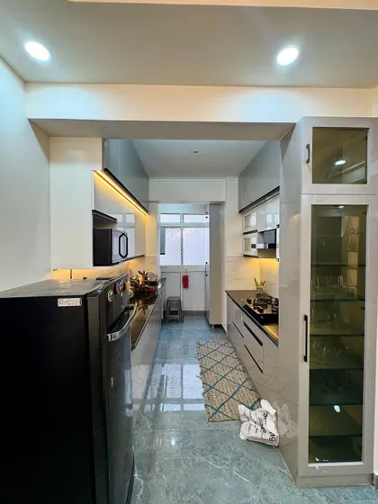 A full view of a completed galley kitchen at Amrapali Princly Estate. The design features dual-tone cabinets, a tall pantry unit, and under-cabinet lighting.