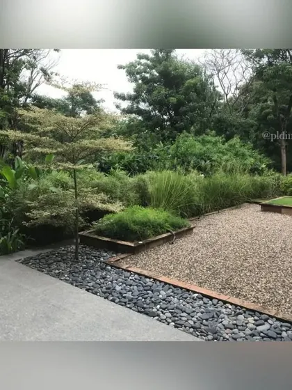 The boundary of the Zen garden is marked by a bed of dark river stones, which provides a strong visual contrast to the light-colored concrete and fine gravel. The layered planting in the background adds softness.