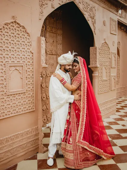An intimate embrace against a stunning architectural backdrop. The groom holds his bride close, her red lehenga a beautiful pop of color against his white sherwani, creating a portrait of royal elegance.
