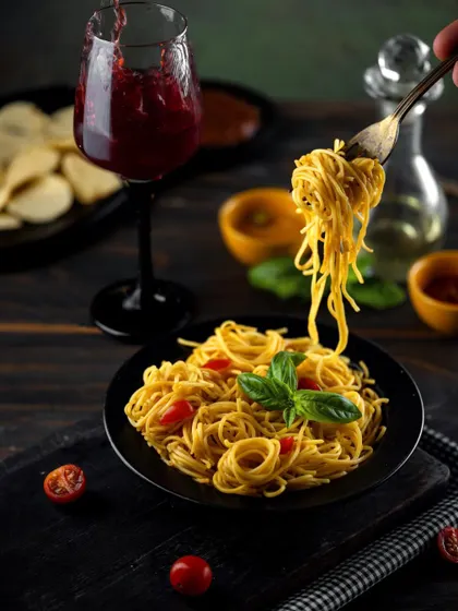 A dark and moody shot of Hakka noodles being lifted with a fork, paired with a glass of red wine. The dramatic lighting highlights the texture of the noodles and creates a sophisticated, indulgent atmosphere.