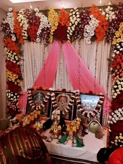 A beautiful backdrop for a home pooja, with deities placed on a decorated platform. The setup is framed with colorful flower arrangements and pink drapes.