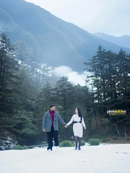 A wide shot of a couple walking hand-in-hand through a snowy path, surrounded by tall pine trees and misty mountains, perfect for a winter pre-wedding shoot.