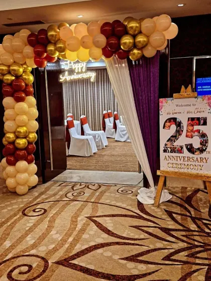 A welcoming entrance for a 25th-anniversary ceremony. The decor includes a balloon arch in cream, maroon, and gold, a 'Happy Anniversary' neon sign, and a personalized welcome easel.