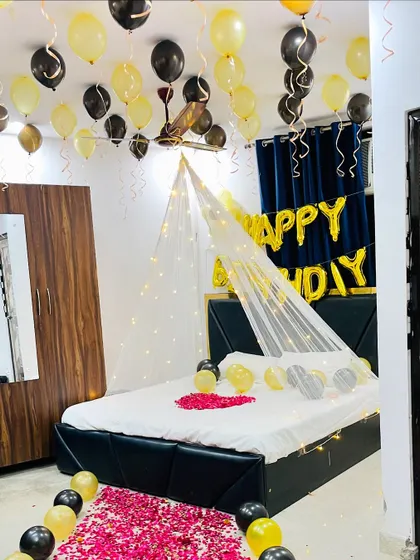 A stylish birthday room decoration in black and gold. The setup includes a fairy light canopy, a rose petal pathway, and 'Happy Birthday' foil balloons against a dark curtain.
