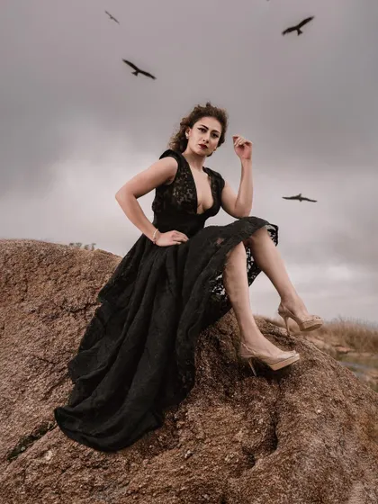 Model in a black lace dress sitting on a rock under a stormy sky with birds flying overhead