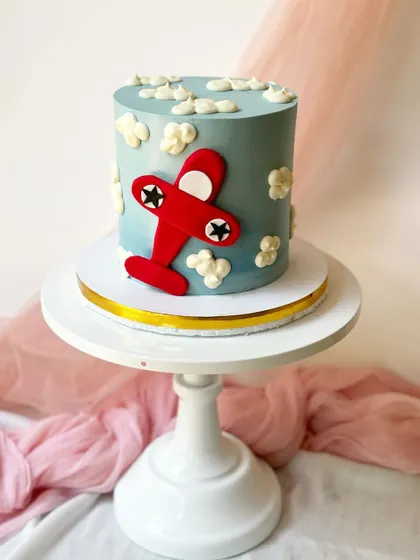 A simple and classic airplane cake. The red fondant plane is set against a sky-blue buttercream background with fluffy white clouds.