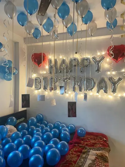 A birthday room decoration with blue and silver balloons. The setup includes 'Happy Birthday' foil balloons with fairy lights and photos hanging from the ceiling.