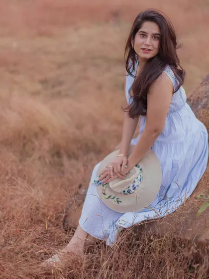 A lovely solo portrait of the bride-to-be sitting in a field of tall grass, holding a hat and looking at the camera with a gentle smile.