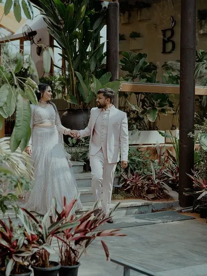 A couple holds hands while walking through a lush, green indoor garden space, dressed in elegant matching outfits.