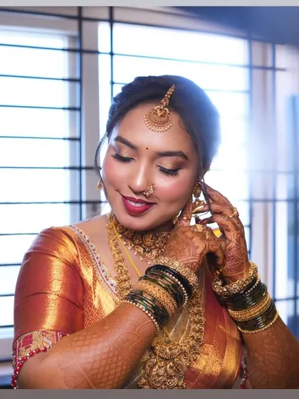 Getting ready for the big moment. This candid shot captures the bride as she puts on her earrings, showing the detailed eye makeup with a golden shimmer and a beautiful berry-toned lip.