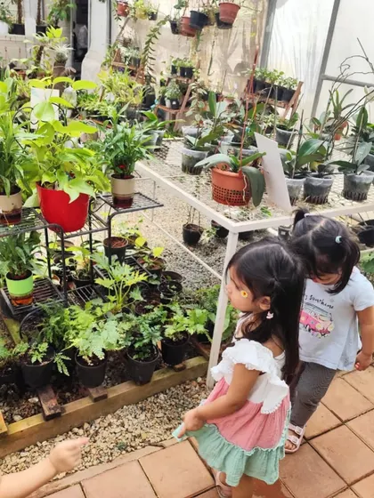 The children navigate through the aisles of the plant nursery, observing the vast variety of plants and flowers.