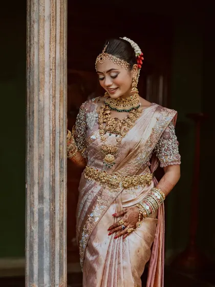 A bride in a pastel-hued saree, adorned with magnificent jewelry, leans against a pillar, the epitome of grace.