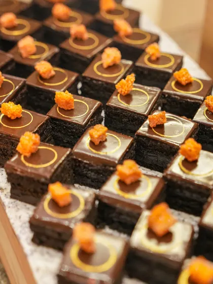 Our opera-style petit fours, featuring layers of cake and cream, topped with a glossy chocolate glaze and a delicate orange garnish. Each square is a perfect, multi-layered bite.