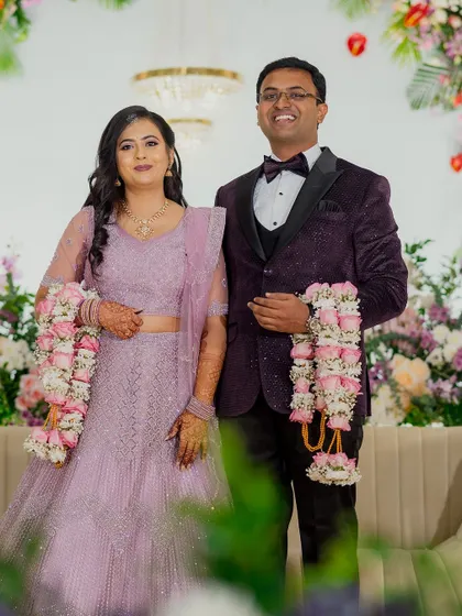 A happy couple at their reception, dressed in beautiful pastel outfits that complement the floral decor. The background features a lush arrangement of pink, white, and lavender flowers.