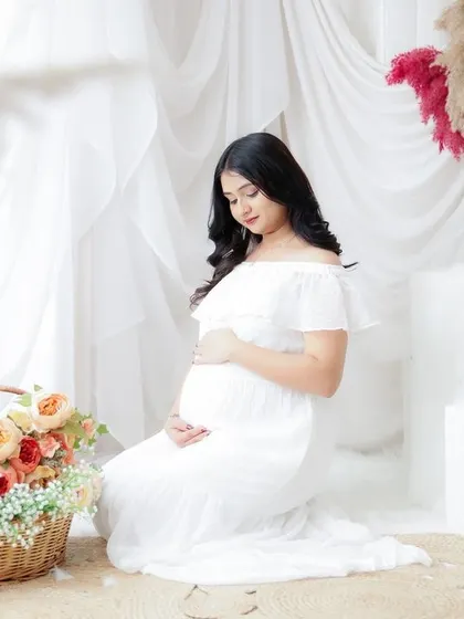 A delicate and serene portrait. The mom-to-be kneels in a simple white dress, surrounded by soft curtains and a basket of flowers.