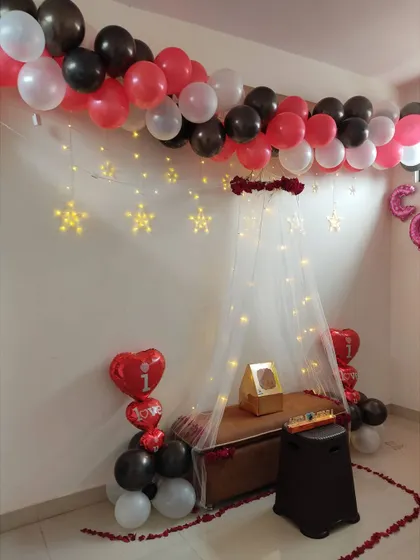 A lovely canopy setup perfect for an anniversary or romantic surprise. The decor includes a balloon garland on the ceiling, star-shaped lights, and a trail of rose petals on the floor.
