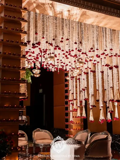A traditional red and white mandap setup inside a grand hall. The decor features pillars adorned with diyas and a ceiling covered in hanging garlands of jasmine and red roses.