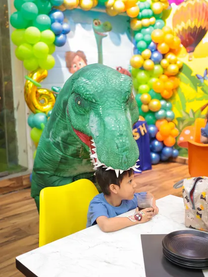 A friendly T-Rex mascot surprises a guest at the dining table during a dinosaur-themed party. It adds a fun and interactive element to the celebration.