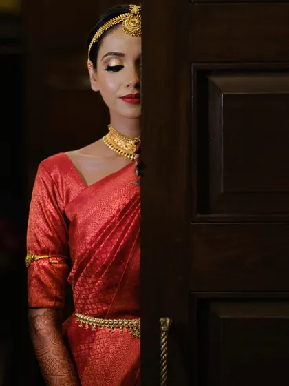 A creative portrait of a South Indian bride, partially hidden behind a door, creating a sense of mystery and anticipation before her grand entrance.