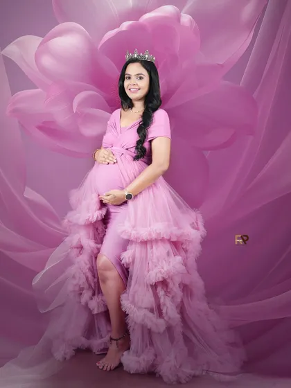 A portrait fit for a queen. The mom-to-be wears a crown and a magnificent pink ruffled gown, set against a dramatic floral backdrop that makes the image pop.