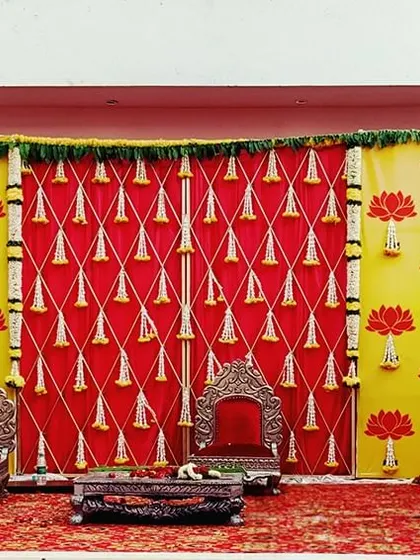 A vibrant and traditional stage setup with a lotus theme. The backdrop uses bright yellow and red fabrics, adorned with lotus cutouts and hanging temple bells for a festive and divine feel.
