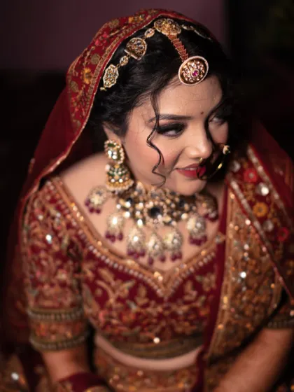 A close-up portrait of a bride, her expression soft and thoughtful. The dark background emphasizes the rich colors of her lehenga and jewelry.