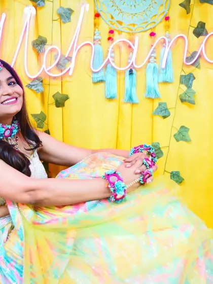 A relaxed and happy portrait of the bride-to-be, seated against the vibrant yellow and pink floral decor of her Mehendi function.