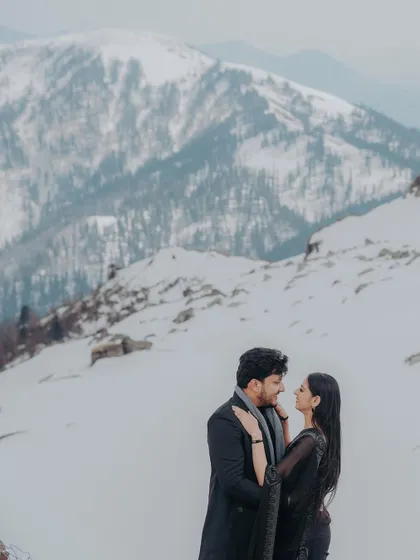 An intimate pre-wedding portrait in a snowy mountain landscape. The couple is dressed in black, creating a beautiful contrast against the white snow and grey skies.