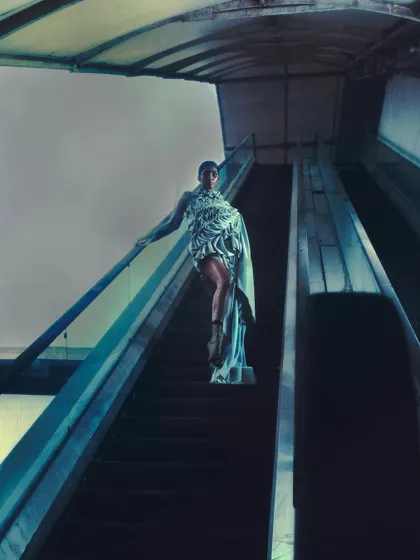 A powerful low-angle shot looking up at the model on an escalator against a stark sky. The composition is monumental, turning a mundane setting into a stage for a high-fashion story.