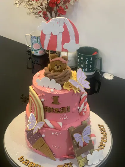 A still photo of the adorable two-tier hot air balloon teddy bear cake. This pink cake is decorated with rainbows, ice cream cones, and butterflies for a whimsical first birthday theme.