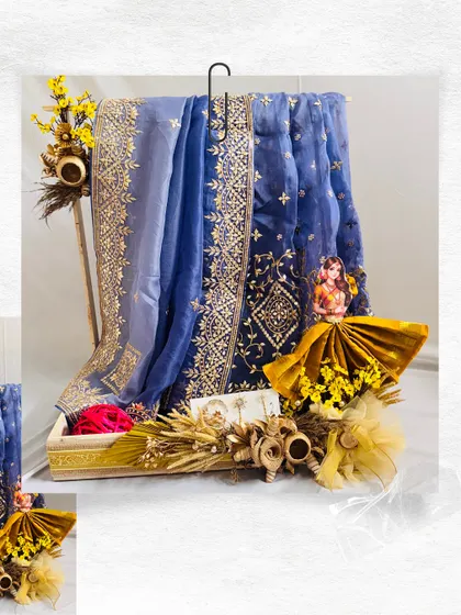 This Teej hamper showcases a stunning blue organza saree with intricate gold embroidery. We complemented it with yellow flowers, dried wheat stalks, and a custom-draped saree doll to create a beautiful and cohesive gift.
