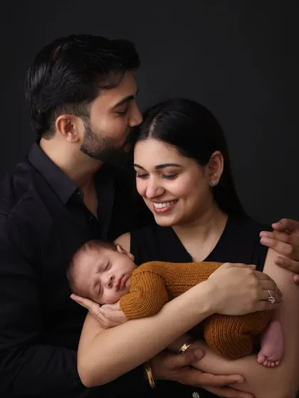 A classic family portrait with a newborn. The parents' loving smiles and the father's gentle kiss on his wife's head create a beautiful frame of family love and unity.